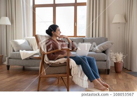 Smiling dreamy Indian woman using laptop, sitting in armchair 108053567