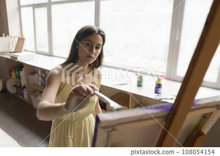 Pretty woman standing near easel embodies creative idea on canvas Pretty woman standing near easel embodies creative idea on canvas 108054154
