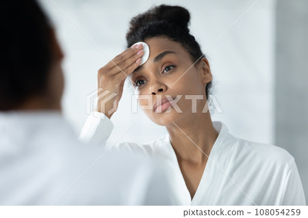 Biracial woman look in bath mirror apply toner with pad 108054259