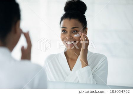 Smiling biracial woman apply face cream in bath 108054762