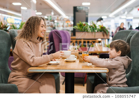 Young curly haired mother and her little son have breakfast or lunch in a cafe, sitting at a table 108055747