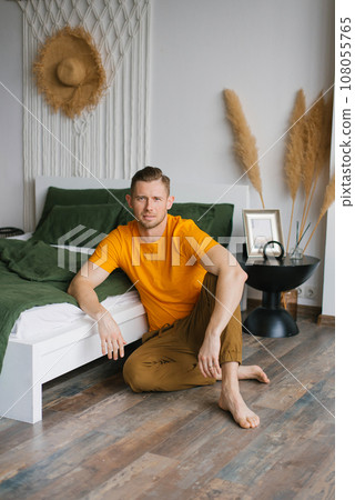 Young man is sitting at home on the floor near the bed and looking at the camera Young man is sitting at home on the floor near the bed and looking at the camera 108055765