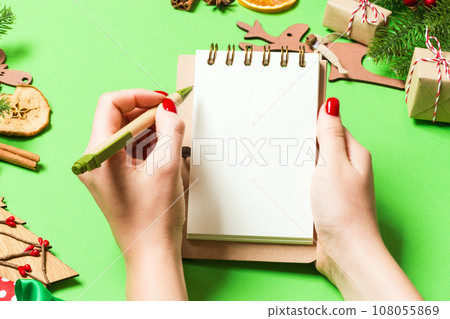 Top view of female hand making some notes in noteebok on green background. New Year decorations and toys. Christmas time concept Top view of female hand making some notes in noteebok on green background. New Year decorations and toys. Christmas time concept 108055869