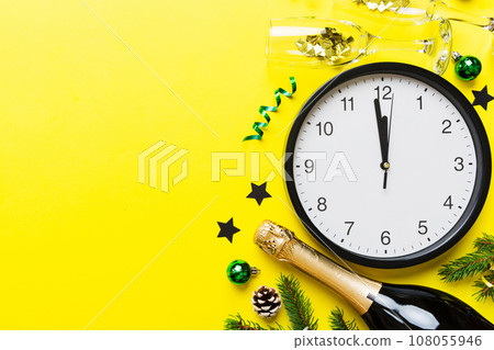 New year celebration concept with a bottle of champagne and two glasses toasting. Christmas gift box, alarm clock and fir tree branch on colored table. Top view Copy space New year celebration concept with a bottle of champagne and two glasses toasting. Christmas gift box, alarm clock and fir tree branch on colored table. Top view Copy space 108055946