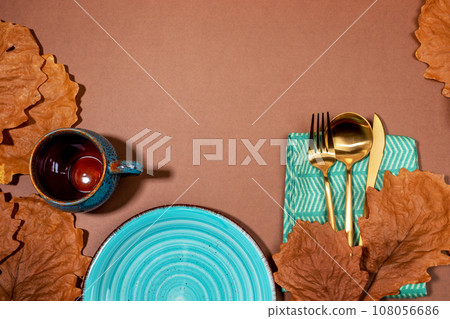 Autumn creative idea of holiday table setting for Halloween or thanksgiving day. Empty turquoise tableware plate and cup, golden cutlery, dry oak leaves on brown background. Top view. Copy space. Autumn creative idea of holiday table setting for Halloween or thanksgiving day. Empty turquoise tableware plate and cup, golden cutlery, dry oak leaves on brown background. Top view. Copy space. 108056686