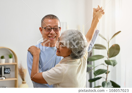Joyful mature grey haired couple dancing in living room while relaxing at home on weekend together Joyful mature grey haired couple dancing in living room while relaxing at home on weekend together 108057023