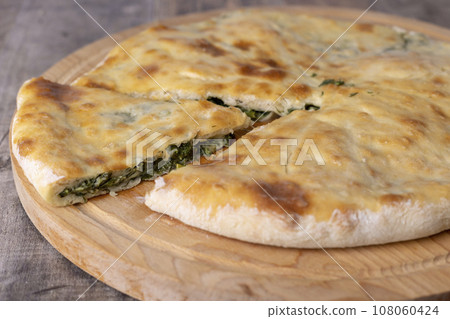 Pie with spinach, herbs and suluguni cheese on a wooden board. Whole pie with cut off piece. Traditional round flat pie with filling. Homemade pastries of Caucasian and Balkarian cuisine.  108060424