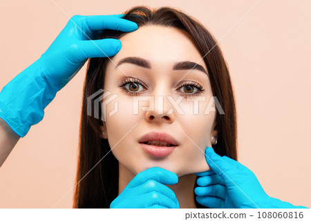 Portrait of a beautiful young woman whose cosmetologists' hands stretch the skin of her face. The concept of massage and rejuvenation Portrait of a beautiful young woman whose cosmetologists' hands stretch the skin of her face. The concept of massage and rejuvenation 108060816