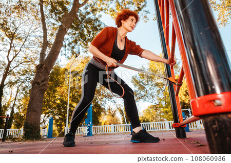 Bottom view of adult smiling Caucasian woman looking in camera does stretching with elastic band on sports playground 108060986