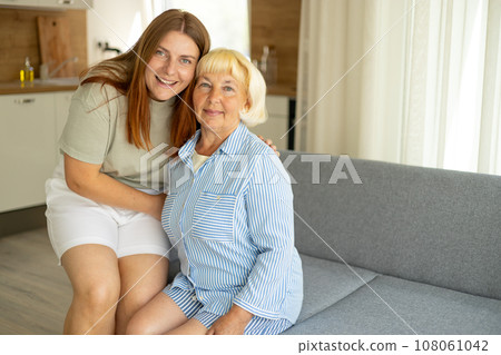 Happy senior woman enjoying in daughter's affection on Mother's day. Smiling mature mother and daughter hugging, sitting on couch at home, looking at camera Happy senior woman enjoying in daughter's affection on Mother's day. Smiling mature mother and daughter hugging, sitting on couch at home, looking at camera 108061042