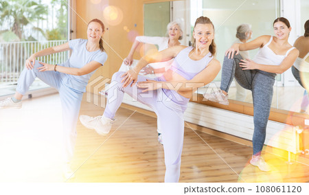 Group of energetic young mature female dancers dancing with knee lift during training together in dance hall 108061120