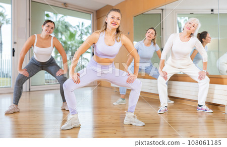Cheerful young mature female dancers in sportswear dancing with legs wide apart during exercising with coach in gym 108061185