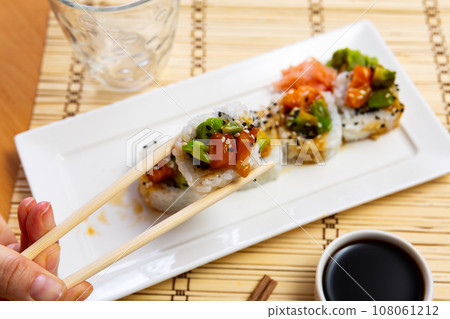 Visitors hands hold piece saketaki roll in chopsticks. Blurred plate with sushi set in background 108061212
