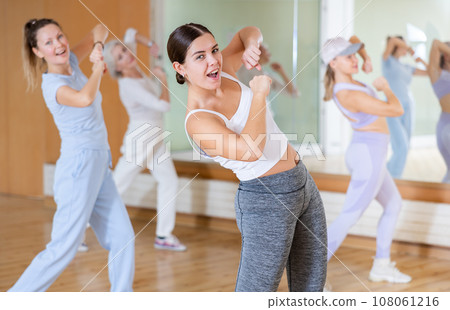 Joyful young mature women in sportswear learning new movements with hands at chest level while dancing together in hall 108061216