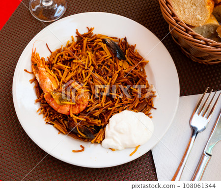 Appetizing traditional Spanish dish made from seafood and noodles served on a plate with aioli sauce on the table in a restaurant Appetizing traditional Spanish dish made from seafood and noodles served on a plate with aioli sauce on the table in a restaurant 108061344