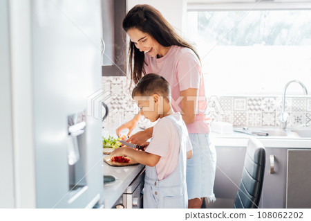 Loving young mother teaching her preschooler son prepare healthy food or salad at home kitchen, caring happy mom cooking together with kid on modern kitchen, boy involved in preparing food with mum. Loving young mother teaching her preschooler son prepare healthy food or salad at home kitchen, caring happy mom cooking together with kid on modern kitchen, boy involved in preparing food with mum. 108062202