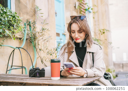 Female traveler chatting, sharing files, resting in cafe terrace table with vintage camera and coffee in reusable cup after city walk. Girl using internet in wifi zone, enjoying coffee and leisure 108062251