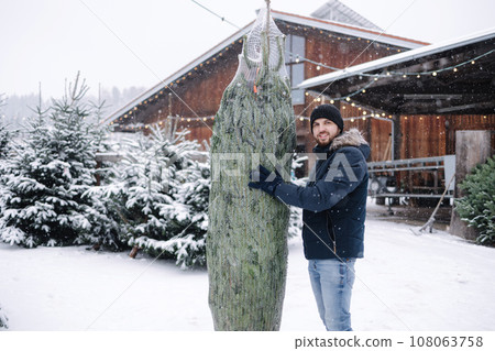 Happy handsome bearded man holds a packed live Christmas tree in outdoor market. Winter snowy day Happy handsome bearded man holds a packed live Christmas tree in outdoor market. Winter snowy day 108063758