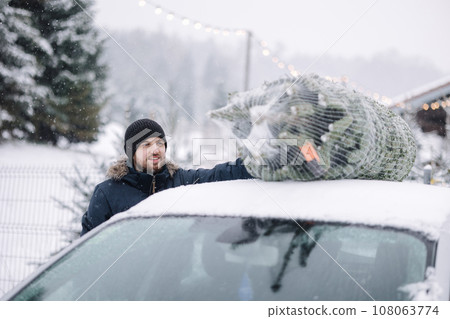 Handsome man tying to put a Christmas tree to the roof of the car to bring it home. Live fir tree delivery Handsome man tying to put a Christmas tree to the roof of the car to bring it home. Live fir tree delivery 108063774