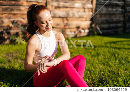 Cheerful sporty female in sportwear smiling looking smart watch screen relaxing sitting on lawn with green grass on sunny summer day. Redhead young sportswoman using checking fitness tracker outdoors. 108064859