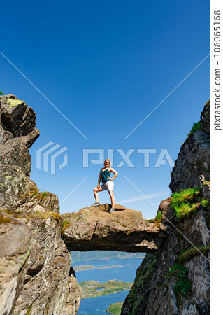 Brave traveler woman standing on hanging stone between rocks. Djevelporten in Norway Lofoten islands 108065168
