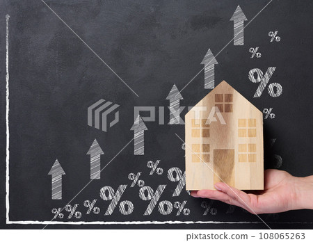 A hand holds a wooden house against the background of a graph with growing indicators, the concept of rising house prices 108065263