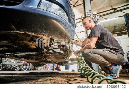 Side view of repairman working in garage, servicing center. Hardworking man holding equipment, changing car wheel, wearing sneakers and dirty clothes. Concept of manual labor. Side view of repairman working in garage, servicing center. Hardworking man holding equipment, changing car wheel, wearing sneakers and dirty clothes. Concept of manual labor. 108065422