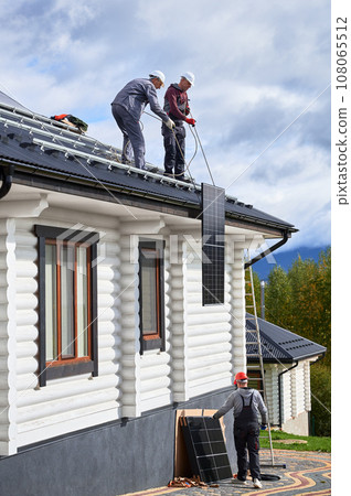 Men technicians installing solar panel system on roof of house. Workers in helmets lifting up photovoltaic solar module with help of ropes outdoors. Concept of alternative and renewable energy. 108065512