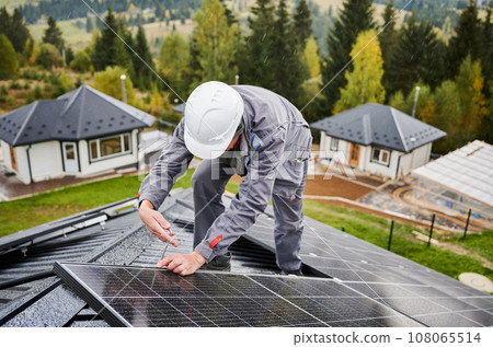 Man electrician mounting photovoltaic solar panels on roof of house. Engineer in helmet installing solar module system with help of hex key. Concept of alternative, renewable energy. Man electrician mounting photovoltaic solar panels on roof of house. Engineer in helmet installing solar module system with help of hex key. Concept of alternative, renewable energy. 108065514