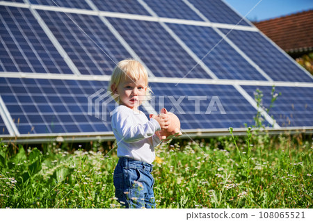 Little cute boy saving money in piggy bank. Small kid learning about saving money for future. Blond child holding piggy bank on background of solar panels at sunny day. Little cute boy saving money in piggy bank. Small kid learning about saving money for future. Blond child holding piggy bank on background of solar panels at sunny day. 108065521