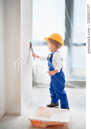 Child construction worker painting wall with paint brush in apartment. Kid in safety helmet and work overalls using paint brush while working at home under renovation. 108065527
