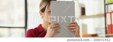Female employee holds clipboard in hands hiding face Female employee holds clipboard in hands hiding face 108072432
