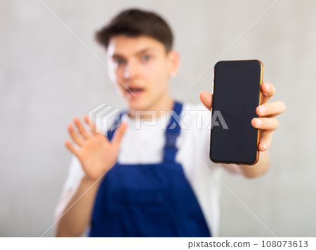 Unfocused man in jumpsuit shows close up empty mobile phone screen and points at device with finger Unfocused man in jumpsuit shows close up empty mobile phone screen and points at device with finger 108073613