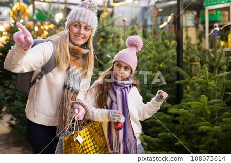 Family of mother and teen daughter having fun on outdoor fair, choosing fir tree for New Year celebration Family of mother and teen daughter having fun on outdoor fair, choosing fir tree for New Year celebration 108073614