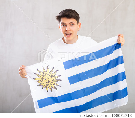 Energized,active male spectator holds national flag of Uruguay in hands and chants popular slogan. 108073718
