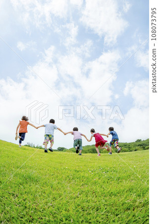 Rear view of elementary school students running up the hill in the park holding hands 108073795