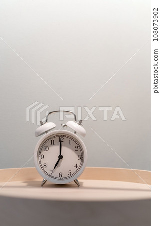 Close-up of a round white alarm clock on a table in the bedroom. The hands on the clock show seven o'clock in the morning, time to get up. Retro alarm clock on the table, vintage tone. space for text 108073902