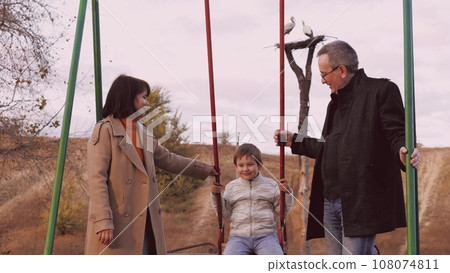 Cheerful parents swing son into air on swing filling moment with laughter. Parents in burst of happiness propel son high into air on swing. Parents laugh and smile swinging son into air on swing Cheerful parents swing son into air on swing filling moment with laughter. Parents in burst of happiness propel son high into air on swing. Parents laugh and smile swinging son into air on swing 108074811