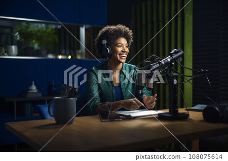 Cheerful smiling female blogger hosting a podcast in the studio. Young African American woman wearing headphones uses laptop, microphone and studio equipment to record live stream for her subscribers. 108075614