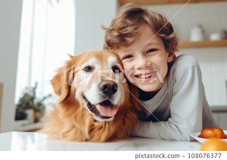 Cheerful little boy is posing with his big shaggy dog at the kitchen table. Funny kid and his pet preparing for breakfast at home. Happy smiling boy and puppy enjoy their time spent together. 108076377