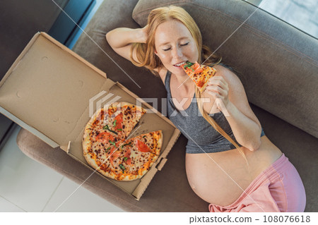 A pregnant woman enjoys a slice of pizza, savoring a moment of indulgence while satisfying her craving for a delightful, comforting treat. Excited Pregnant Young Lady Enjoying Pizza Holding Biting 108076618