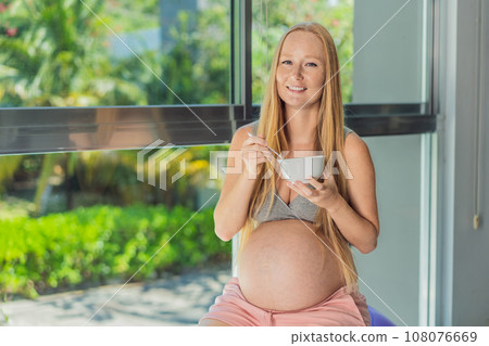 An expecting mother gracefully has breakfast on a fitness ball, illustrating the comfort it provides during pregnancy when traditional chairs may be less accommodating 108076669