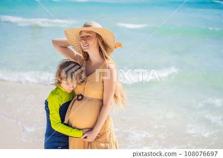 A radiant pregnant mother and her excited son share a tender moment on a serene, snow-white beach, celebrating family love amidst nature's beauty A radiant pregnant mother and her excited son share a tender moment on a serene, snow-white beach, celebrating family love amidst nature's beauty 108076697
