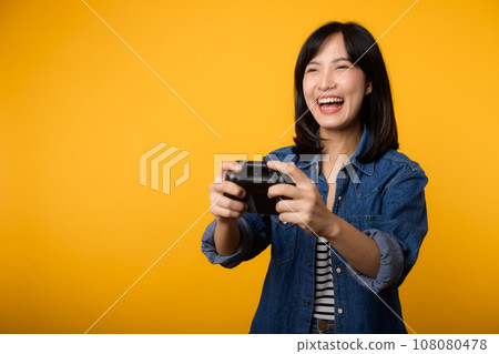 Portrait young asian woman with happy success smile wearing denim clothes holding joystick controller and playing video game. Fun and relax hobby entertainment lifestyle concept. 108080478