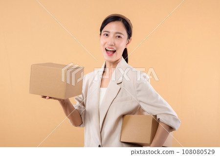 portrait of happy successful confident young asian business woman wearing white jacket holding parcel box isolated on beige studio background, Delivery courier and shipping service concept. 108080529