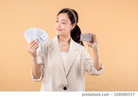 portrait of happy successful confident young asian business woman wearing white jacket holding cash money dollars and credit card standing over beige background. millionaire business, shopping concept 108080557