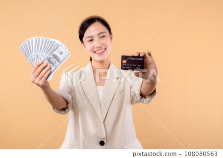 portrait of happy successful confident young asian business woman wearing white jacket holding cash money dollars and credit card standing over beige background. millionaire business, shopping concept portrait of happy successful confident young asian business woman wearing white jacket holding cash money dollars and credit card standing over beige background. millionaire business, shopping concept 108080563
