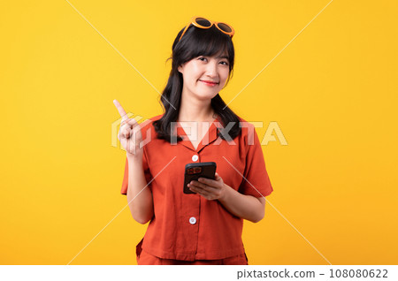 Portrait beautiful young asian woman happy smile dressed in orange clothes with smartphone and pointing finger hand gesture to free space isolated on yellow studio background. app smartphone concept Portrait beautiful young asian woman happy smile dressed in orange clothes with smartphone and pointing finger hand gesture to free space isolated on yellow studio background. app smartphone concept 108080622