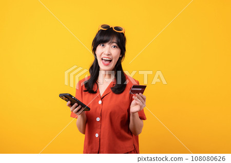 Portrait young beautiful asian woman happy smile dressed in orange clothes using credit card purchase shopping online on smartphone isolated on yellow background. Pay and purchase payment concept. Portrait young beautiful asian woman happy smile dressed in orange clothes using credit card purchase shopping online on smartphone isolated on yellow background. Pay and purchase payment concept. 108080626