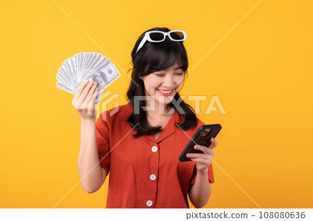 Portrait young beautiful asian woman happy smile dressed in orange clothes showing cash money and smartphone isolated on yellow background. Pay and purchase shopping online payment concept. Portrait young beautiful asian woman happy smile dressed in orange clothes showing cash money and smartphone isolated on yellow background. Pay and purchase shopping online payment concept. 108080636